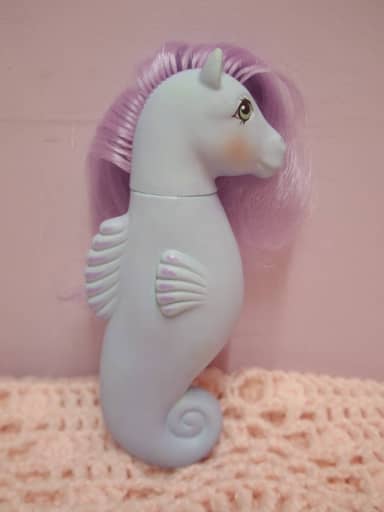 Poney de mer bleu pâle à queue en spirale et crinière mauve, posé sur un tissu rose.