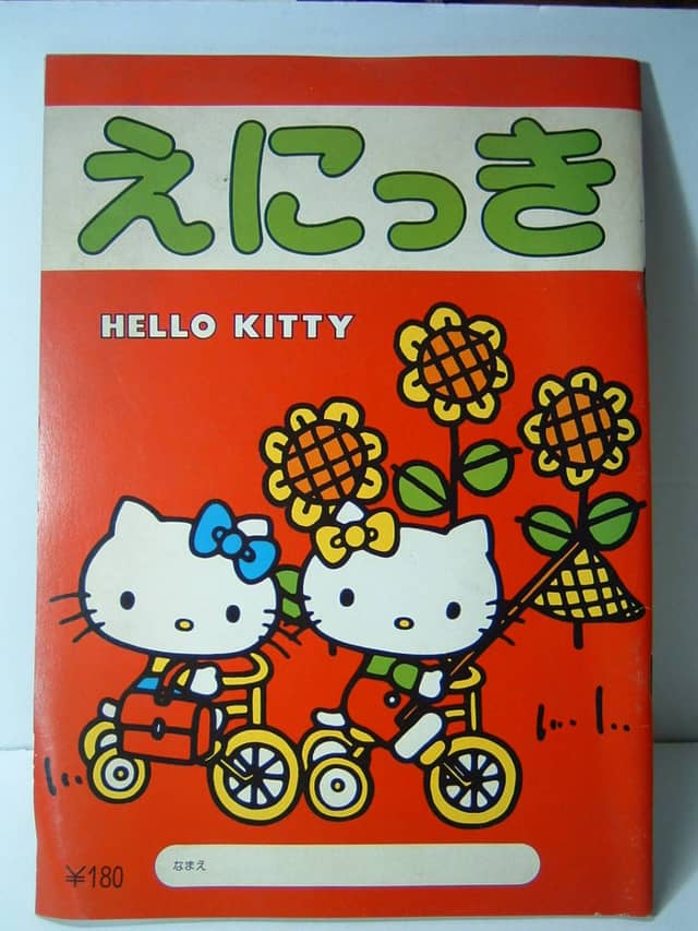 Orange cover of an “えにっき” notebook showing two Hello Kittys cycling among sunflowers.
