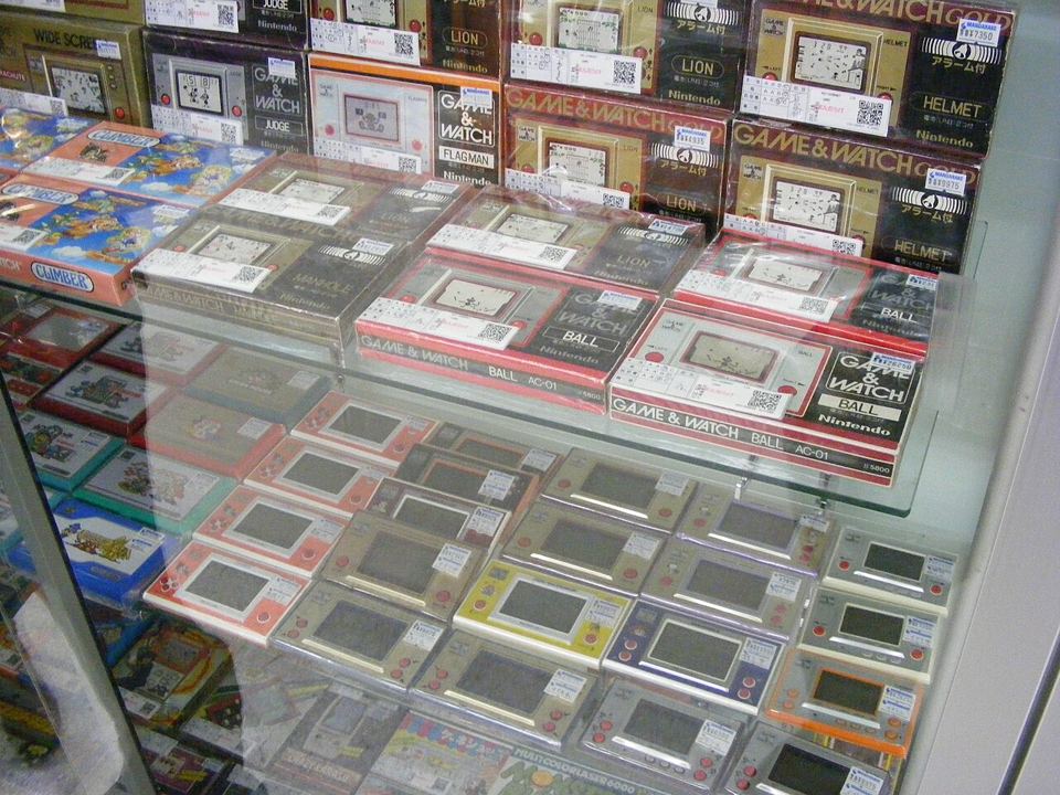 Vitrine d'une boutique d'occasion remplie de Game & Watch : consoles en boîte (Ball, Lion, Helmet, Flagman) en haut, consoles seules étiquetées en dessous.